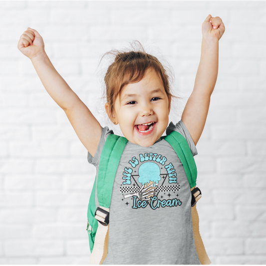 A cheerful young child with a big smile and raised arms, wearing a heather-gray graphic tee with the slogan "Life is Better with Ice Cream" and an illustration of an ice cream cone. The child is also wearing a green and beige backpack, standing against a white brick background.