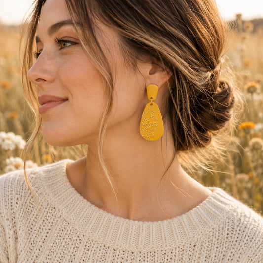 Tea-Shirt Shoppe mustard yellow teardrop dangle earrings with embossed botanical leaf pattern worn by a woman.