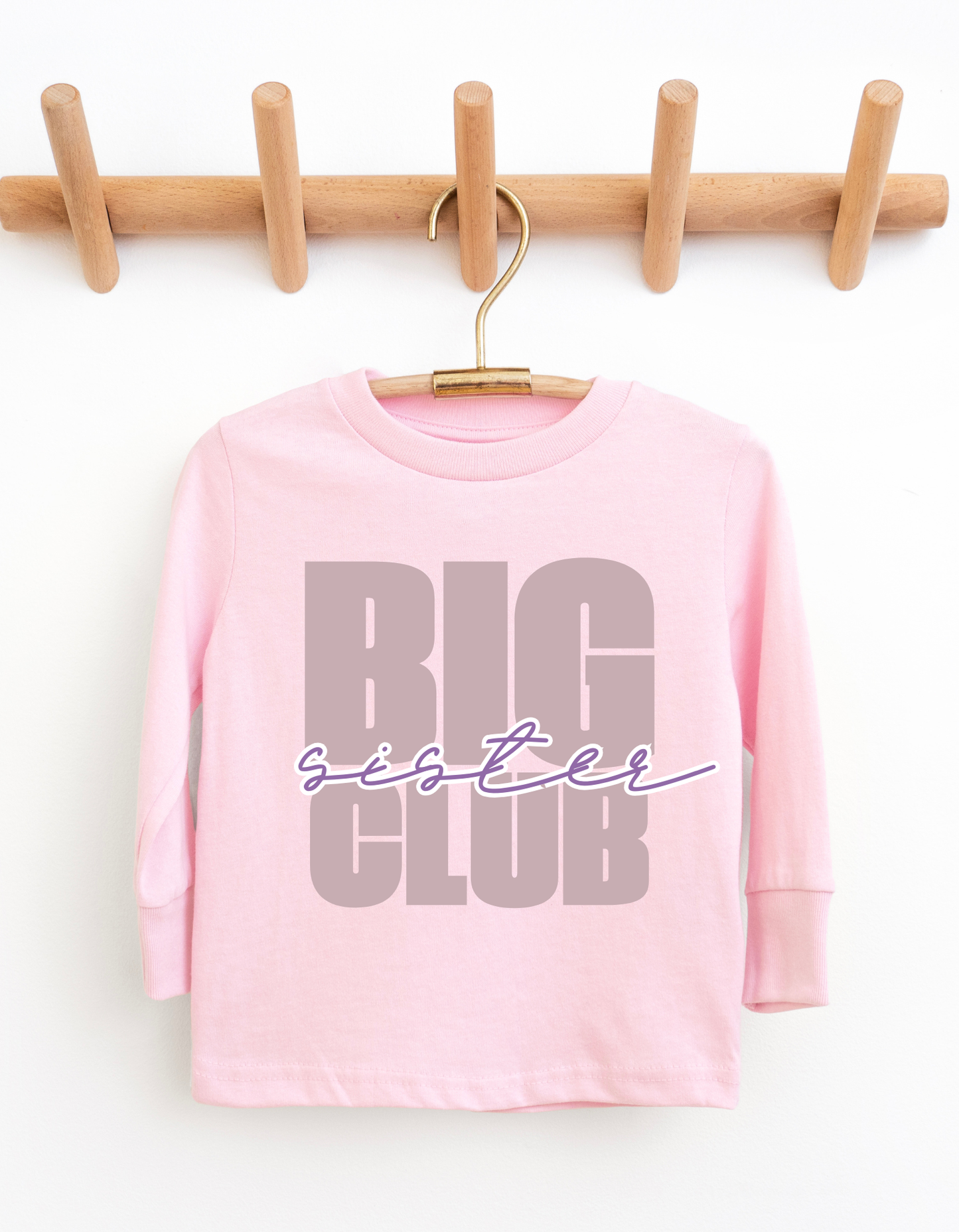 Pink long-sleeve shirt with 'Big Sister Club' text on a wooden hanger against a white background