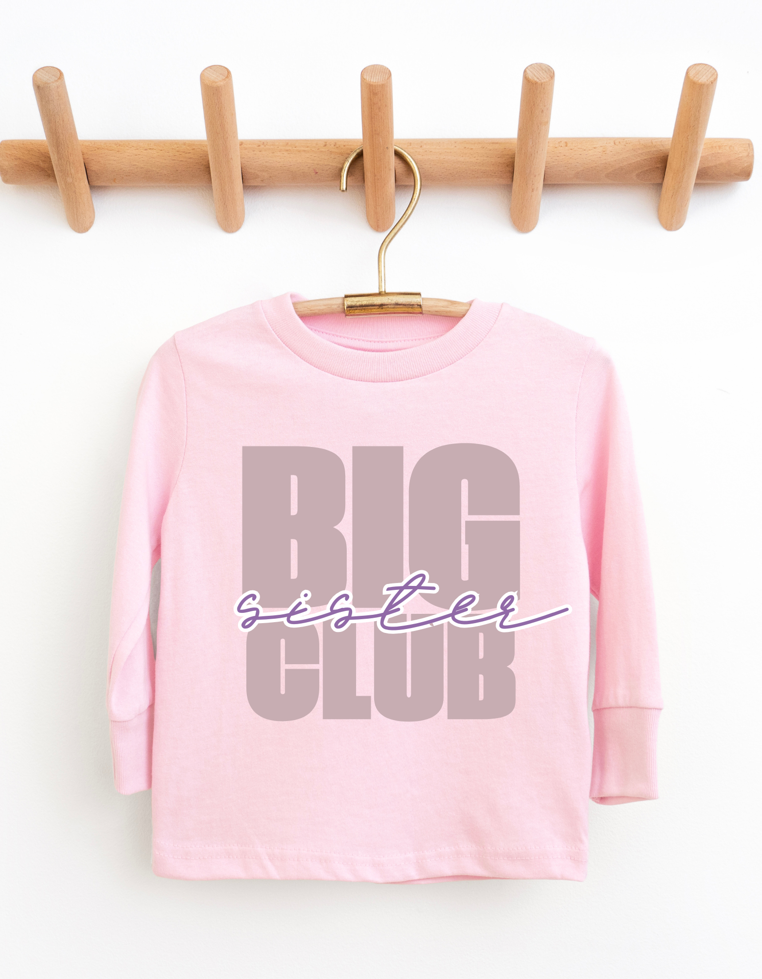 Pink long-sleeve shirt with 'Big Sister Club' text on a wooden hanger against a white background