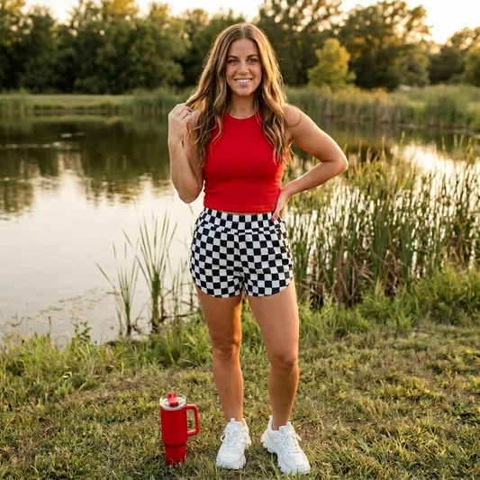 Woman in red top and checkered shorts standing by a lake with a red mug on the ground.