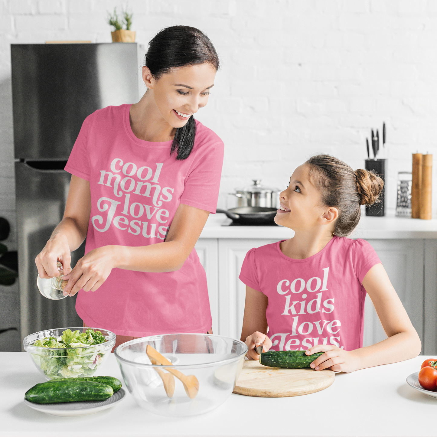 Woman and child in pink 'cool moms love Jesus' shirts preparing food in a kitchen.