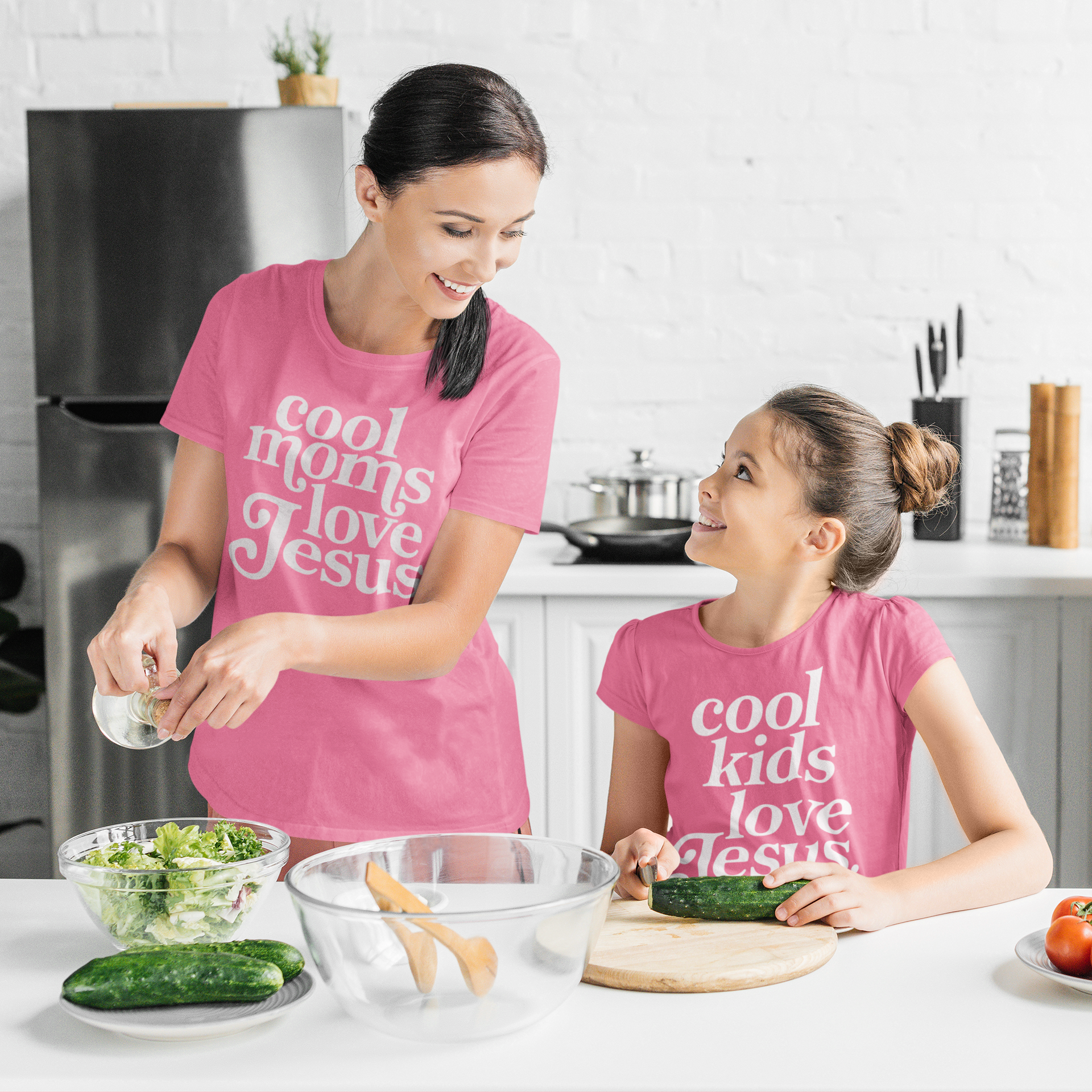 Woman and child in pink 'cool moms love Jesus' shirts preparing food in a kitchen.