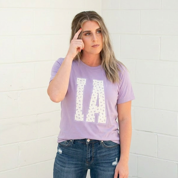 Woman wearing a purple t-shirt with 'IA' and blue jeans against a white brick wall.