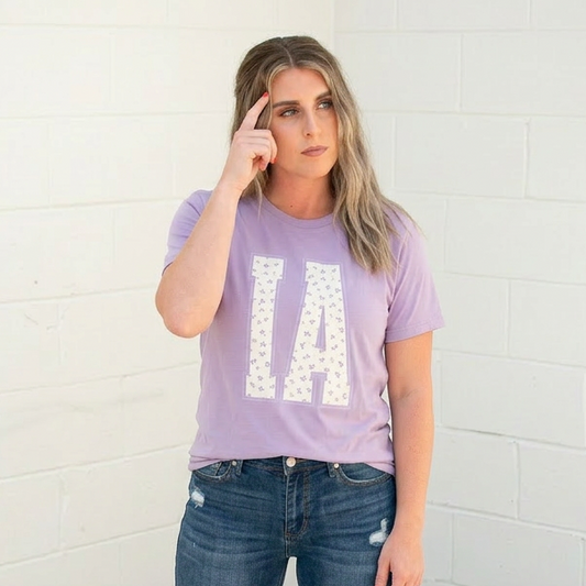 Woman wearing a purple t-shirt with 'IA' and blue jeans against a white brick wall.