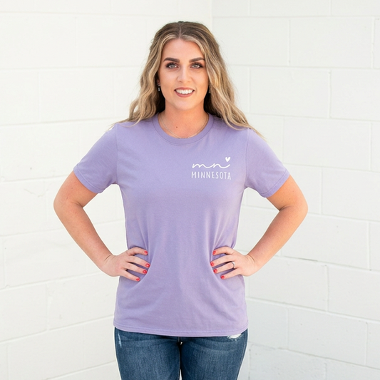 Woman wearing a purple Minnesota t-shirt with text and jeans standing against a white brick wall.