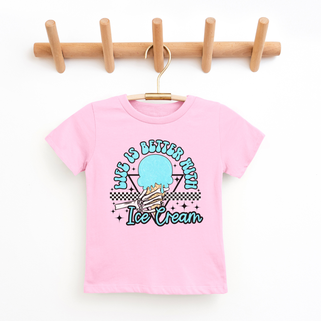 A pink kids' graphic t-shirt displayed on a wooden hanger against a clean, white background. The shirt features a colorful design in the center with the phrase "Life is Better with Ice Cream" in playful lettering, encircling a stylized image of an ice cream cone with a checkered motif at the bottom. The t-shirt has a classic crew neckline and short sleeves, suggesting a comfortable and casual style. The hanger is mounted on a wooden rack with four pegs, providing a natural and simple presentation.