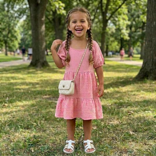 Young girl in a pink dress standing in a park with trees and grass in the background.