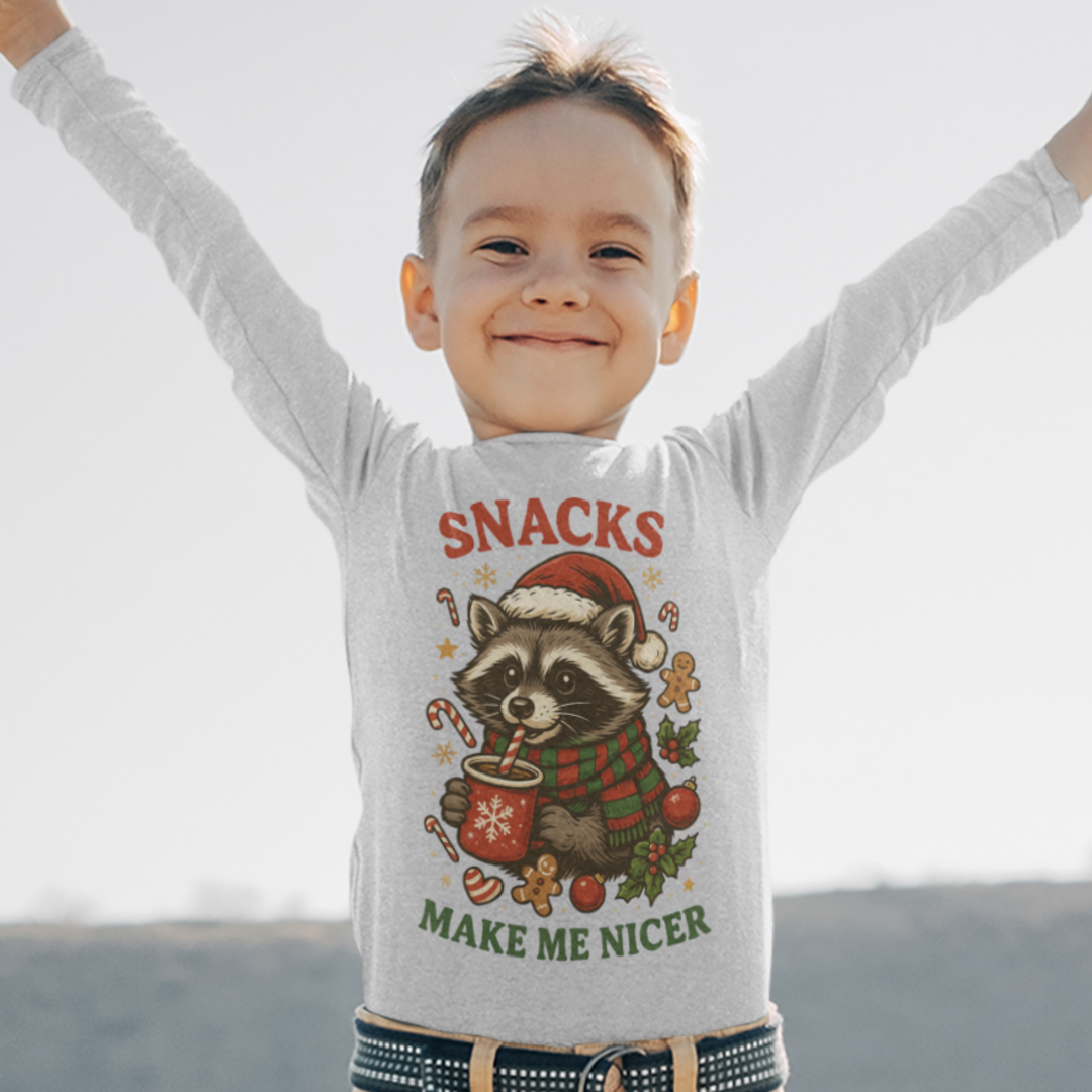 Smiling child with arms raised wearing a gray long-sleeve kids Christmas shirt featuring a raccoon in a Santa hat, red and green scarf, cocoa mug, candy canes, gingerbread cookies, and the festive text “Snacks Make Me Nicer.”