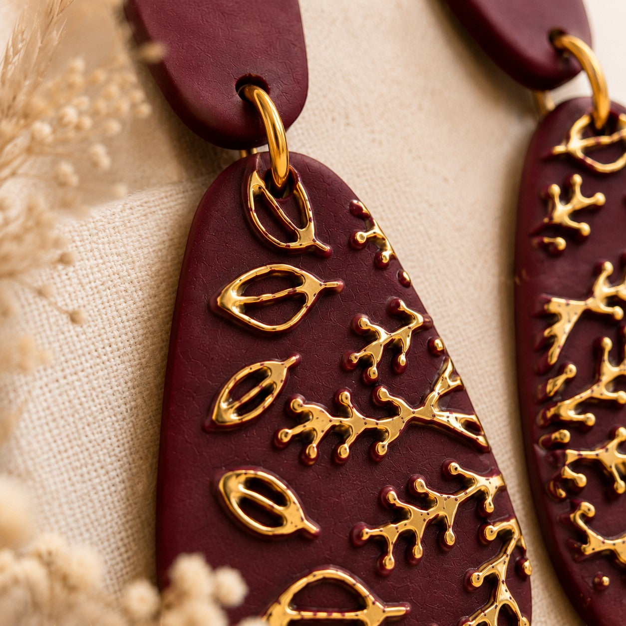 Tea-Shirt Shoppe burgundy teardrop dangle earrings with gold botanical leaf and branch details on a textured background.