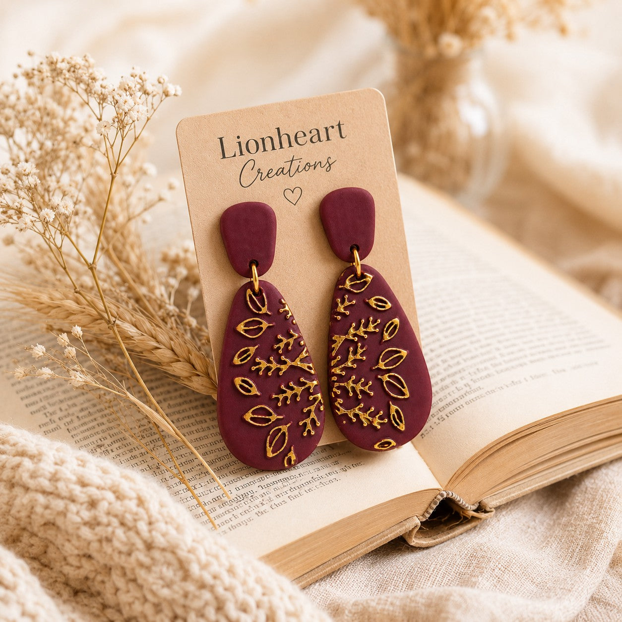 Tea-Shirt Shoppe burgundy teardrop dangle earrings with gold botanical leaf details on a display card by a book.