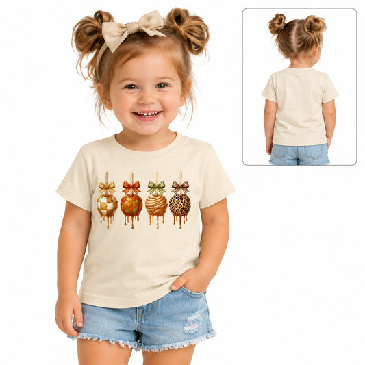 Tea-Shirt Shoppe cream toddler graphic tee featuring four caramel apples with bows, worn by a smiling young girl.