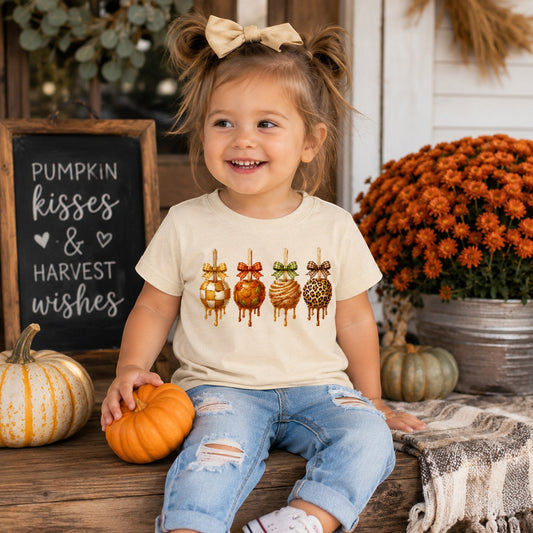 Tea-Shirt Shoppe cream graphic tee featuring four caramel apples with bows, worn by a toddler in a fall setting.