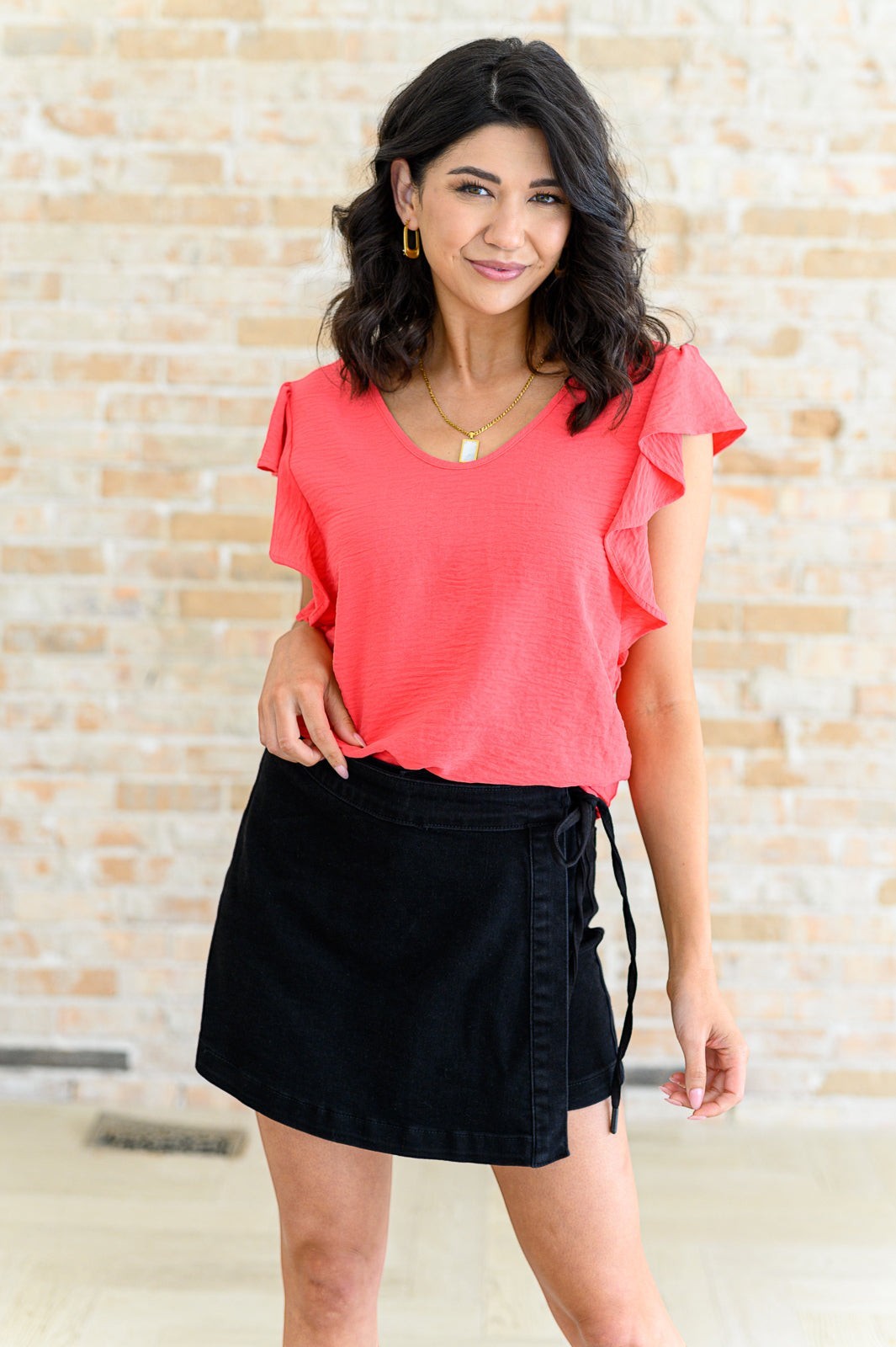 Tea-Shirt Shoppe coral crinkle woven top with flutter sleeves and round neckline worn by a model with a black skirt