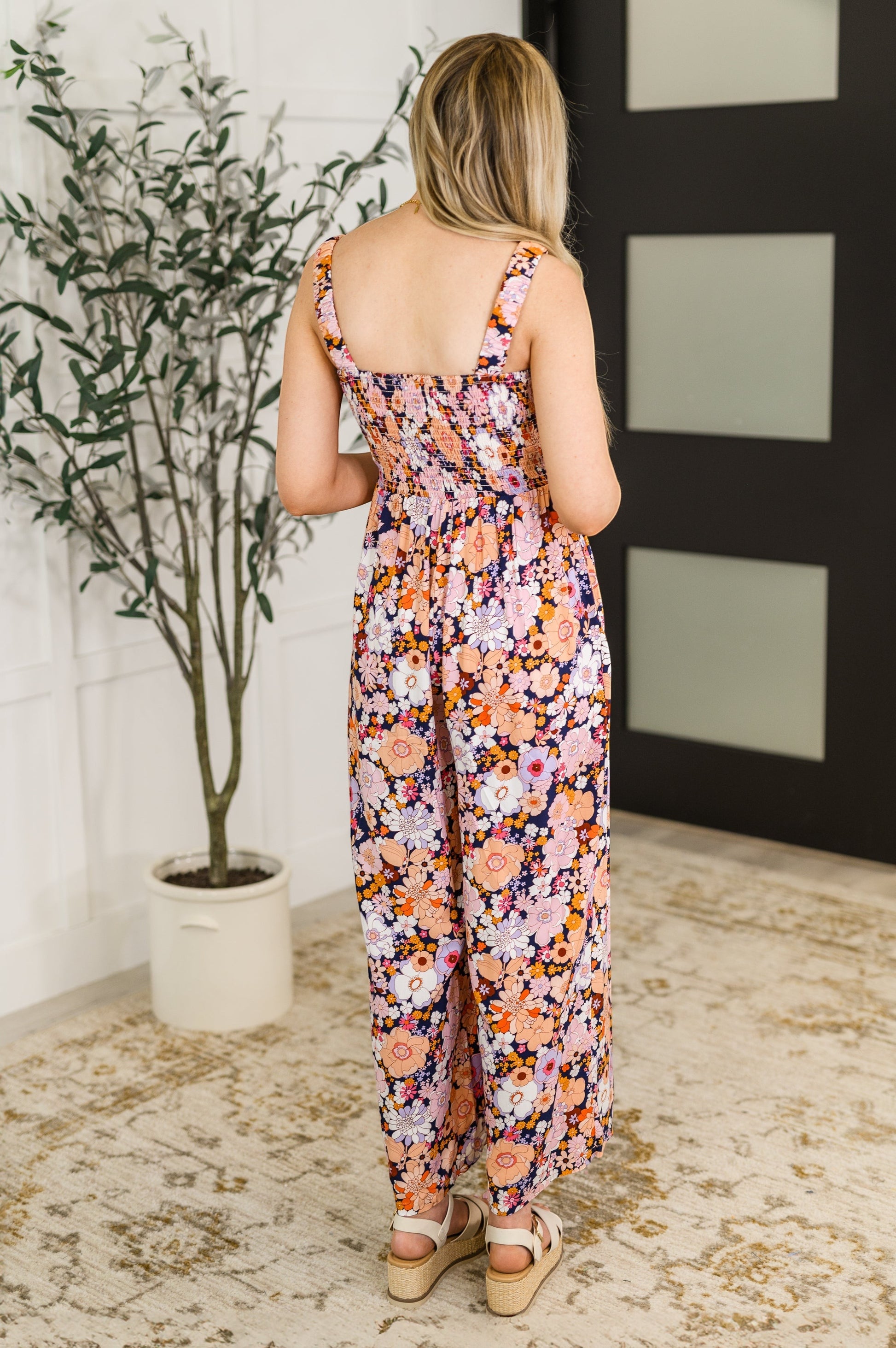 Tea-Shirt Shoppe floral jumpsuit with smocked bodice, square neckline, and wide legs, worn by a model from the back.
