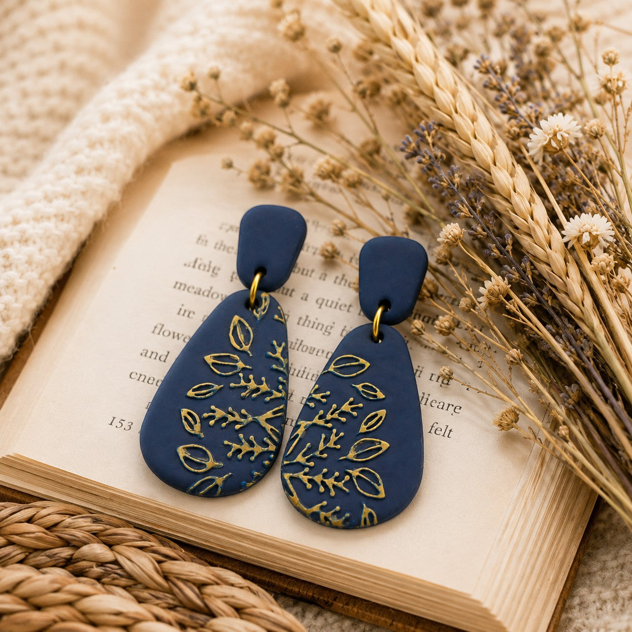 Tea-Shirt Shoppe navy blue polymer clay teardrop earrings with embossed gold botanical details on an open book.