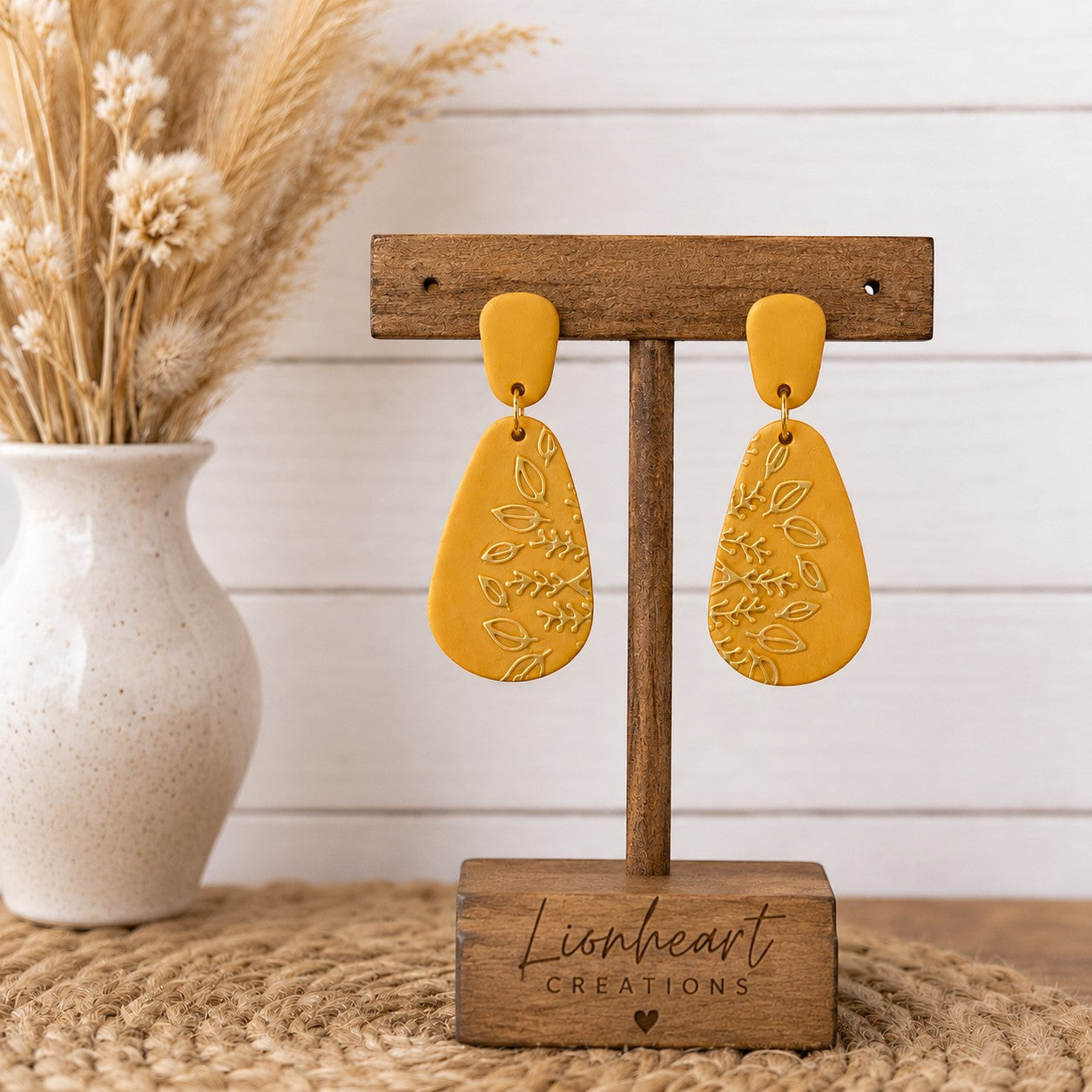 Tea-Shirt Shoppe mustard yellow teardrop dangle earrings with embossed botanical leaf patterns on a wooden stand.