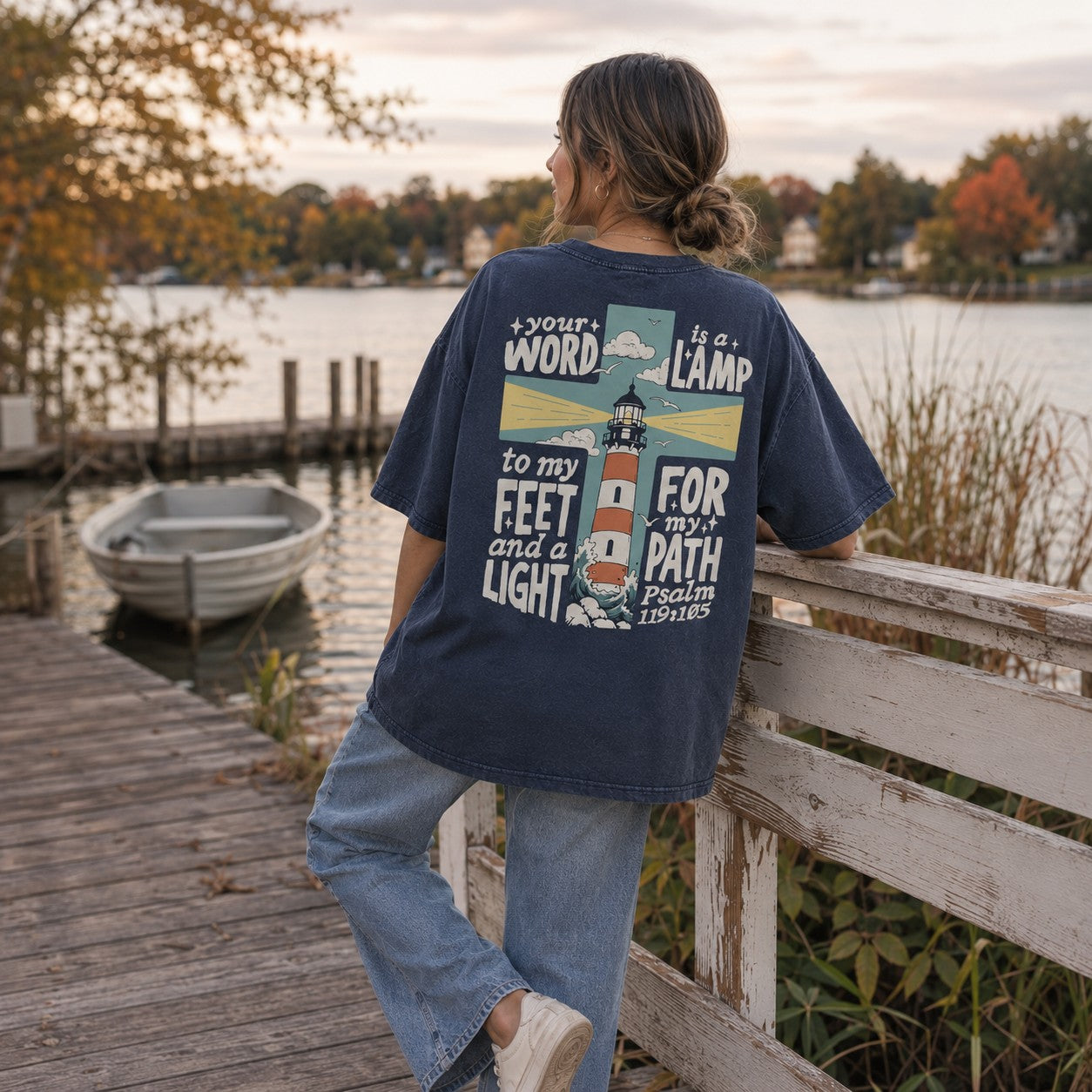 Tea-Shirt Shoppe navy graphic tee with a lighthouse and scripture print on the back, worn by a woman on a dock.