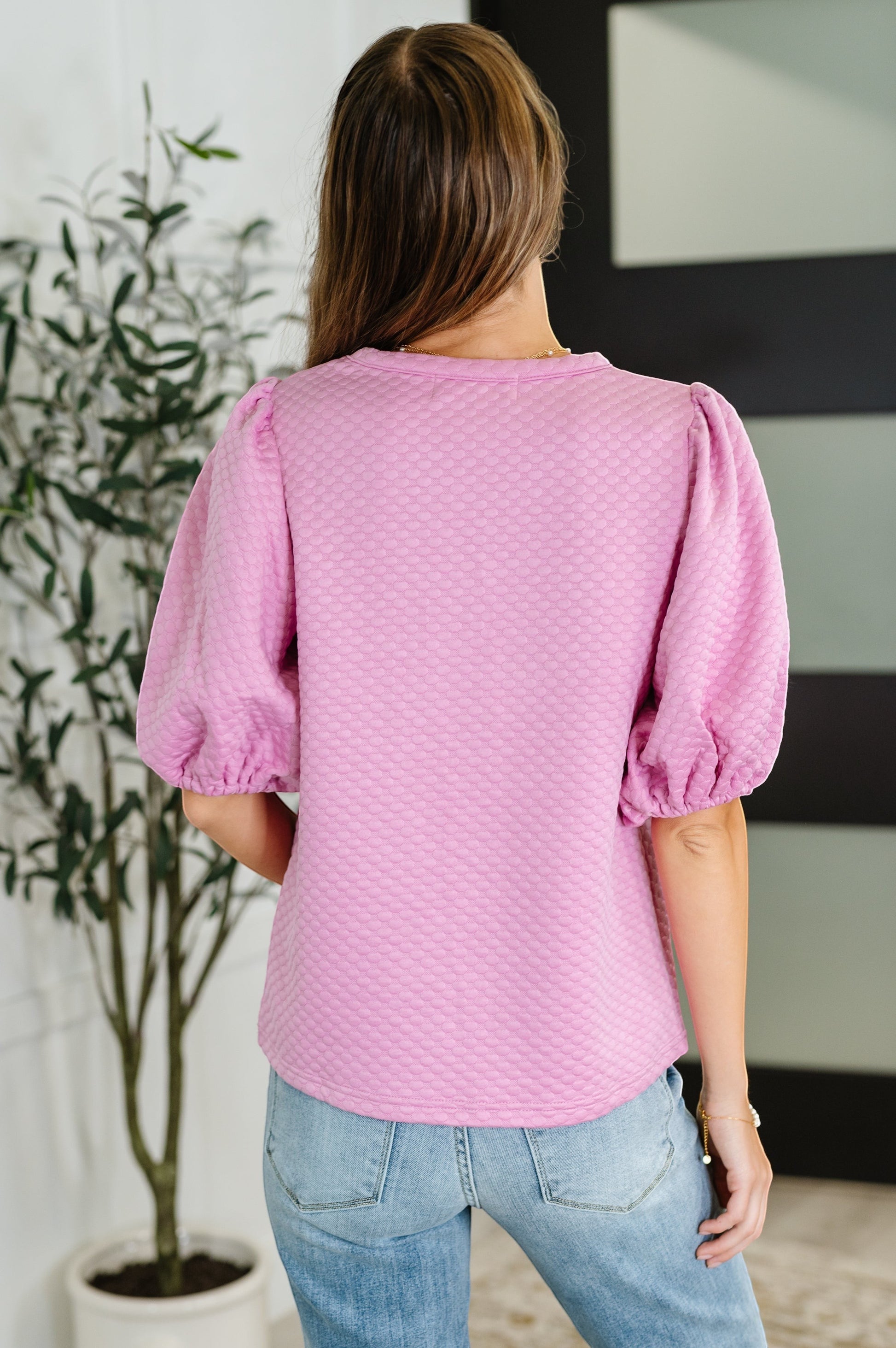 Tea-Shirt Shoppe pink textured blouse with puff sleeves and round neckline worn by a model, back view.