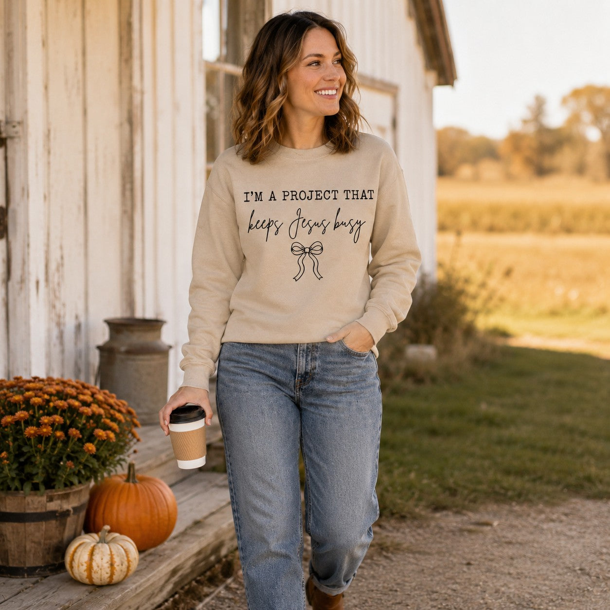 Tea-Shirt Shoppe sand crewneck sweatshirt with Keeps Jesus Busy graphic, worn by a woman on a rustic porch.