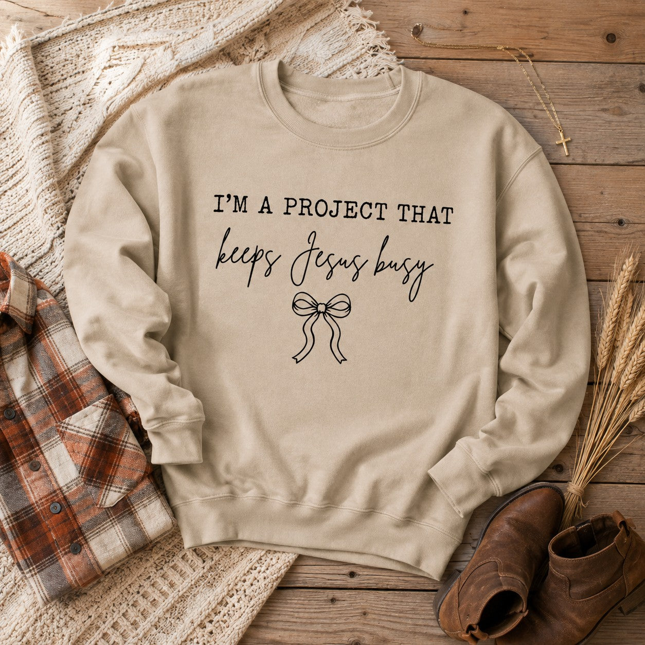 Tea-Shirt Shoppe sand-colored crewneck sweatshirt with I'm a Project That Keeps Jesus Busy text and a bow graphic.