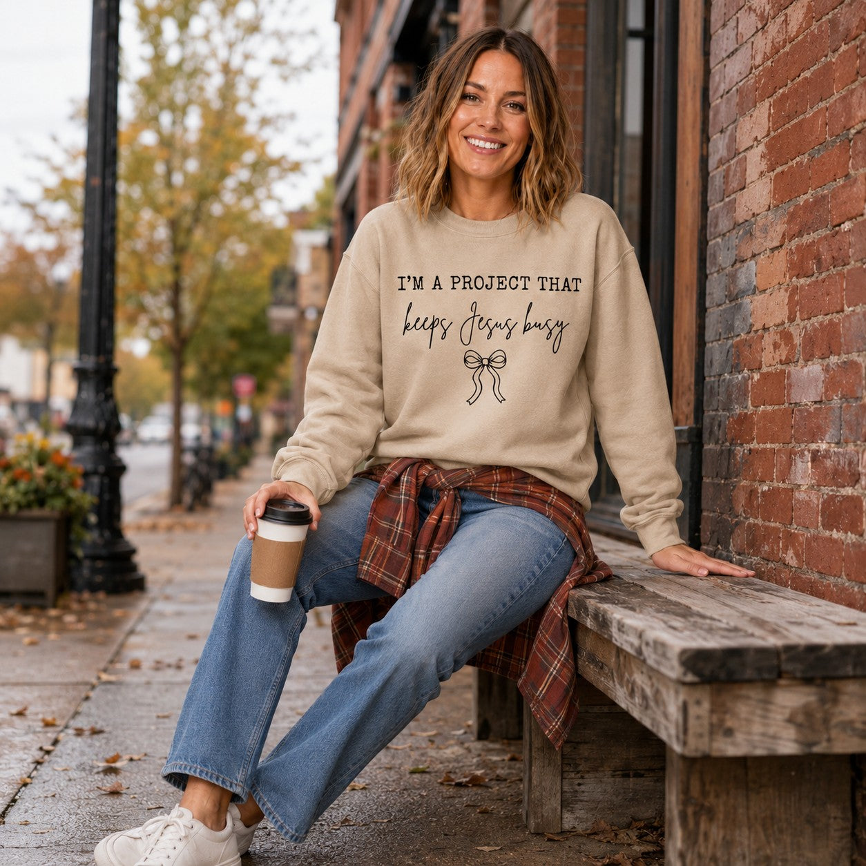 Tea-Shirt Shoppe sand crewneck sweatshirt with I'm a Project That Keeps Jesus Busy text and bow graphic on a woman