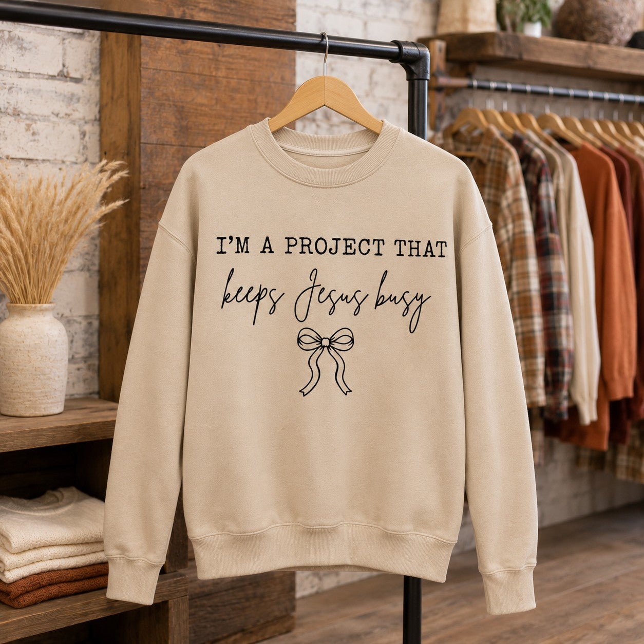 Tea-Shirt Shoppe sand-colored crewneck sweatshirt with I'm a Project That Keeps Jesus Busy text and a bow graphic.