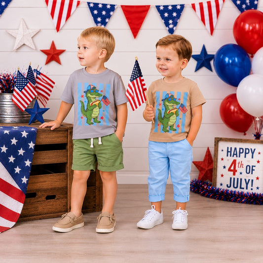 Tea Shirt Shoppe Wholesale: Two young boys in graphic tees featuring a cartoon alligator holding a hot dog and American flags. One tee is grey, the other tan.