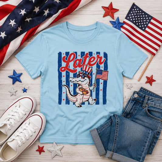 Tea Shirt Shoppe Wholesale light blue graphic tee with a patriotic alligator wearing Uncle Sam's hat and sunglasses, holding an American flag and a hot dog.