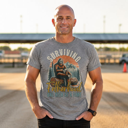 Tea Shirt Shoppe Wholesale grey graphic tee with Bigfoot holding a beer, "Surviving Fatherhood One Beer at a Time" text.