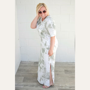 Smiling woman with sunglasses wearing an olive and white maxi dress with slight side slit.