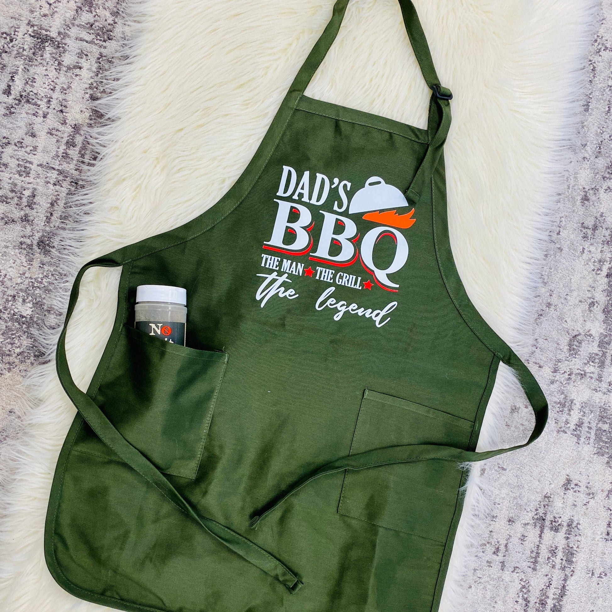 Get your dad an apron he'll love! This green apron is perfect for a sleek masculine look. On the front is the graphic that reads the following, "Dad's BBQ The Man, The Grill, The Legend" With a grill top and flames. 